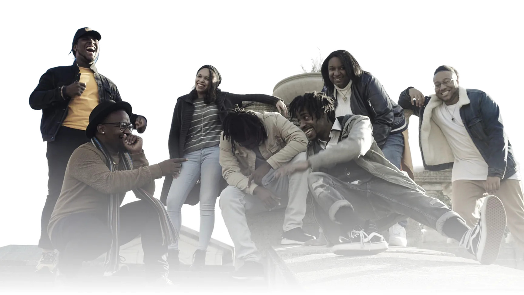 A group of six young adults outdoors smiling and laughing together, with three people standing and three sitting or crouching on a stone structure.